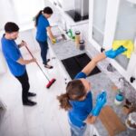 Two individuals cleaning a kitchen, one using a mop and the other holding a dustpan, ensuring a tidy environment.