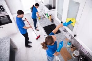 Two individuals cleaning a kitchen, one using a mop and the other holding a dustpan, ensuring a tidy environment.