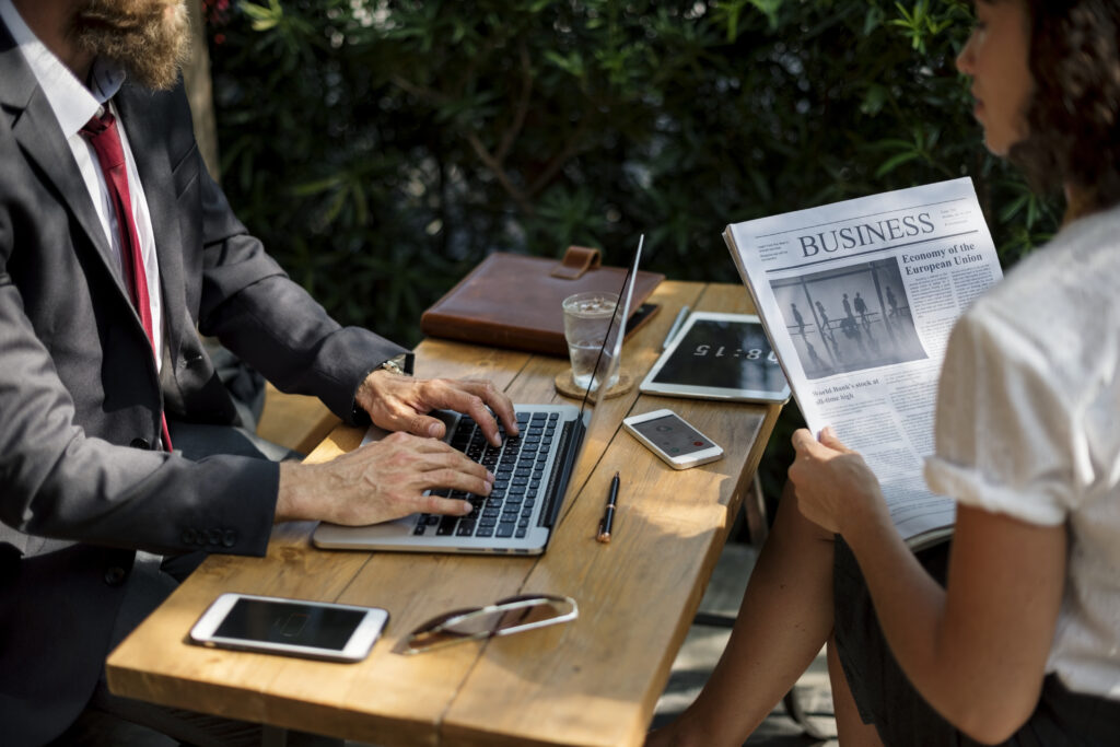 Two professionals sitting at an outdoor table, one typing on a laptop while the other reads a business newspaper, symbolizing the intersection of traditional print media and digital news distribution.