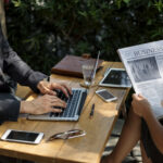 Two professionals sitting at an outdoor table, one typing on a laptop while the other reads a business newspaper, symbolizing the intersection of traditional print media and digital news distribution.