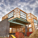 A low-angle view of a residential building site demonstrating the house construction process in the UK, featuring an exposed timber frame on the upper level and dark brickwork being installed on the ground floor under a cloudy sky.