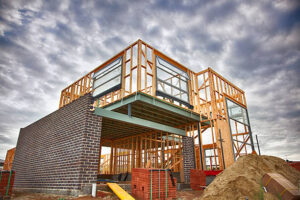 A low-angle view of a residential building site demonstrating the house construction process in the UK, featuring an exposed timber frame on the upper level and dark brickwork being installed on the ground floor under a cloudy sky.
