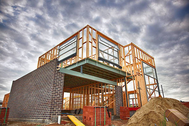 A low-angle view of a residential building site demonstrating the house construction process in the UK, featuring an exposed timber frame on the upper level and dark brickwork being installed on the ground floor under a cloudy sky.