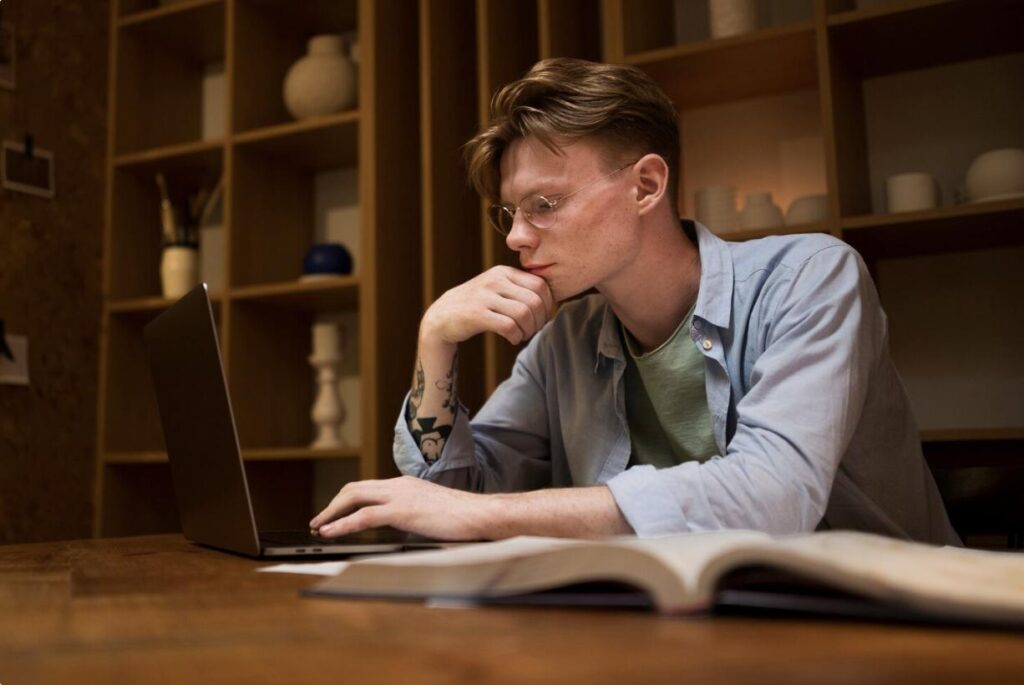 A young man sitting at a desk at home, focused on a laptop screen while studying for his NREMT cognitive exam.