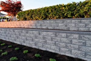 Retaining walls in construction: a two-tier wall built with grey textured concrete blocks in a residential backyard, featuring a green hedge and a red Japanese maple tree.