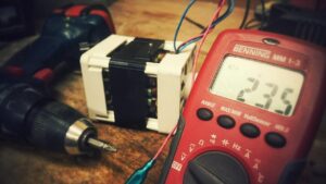 A close-up shot showing a technician using a red digital multimeter to test the voltage of an electrical component on a workbench