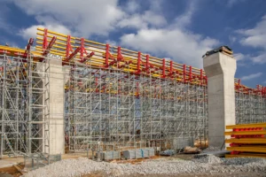 A large-scale construction site featuring complex scaffolding and formwork systems representing temporary works in building construction between concrete piers.