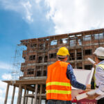Two site engineers in safety vests and hard hats analyzing a digital-ready blueprint against a multi-story building frame, illustrating smart construction trends 2026 in project management.