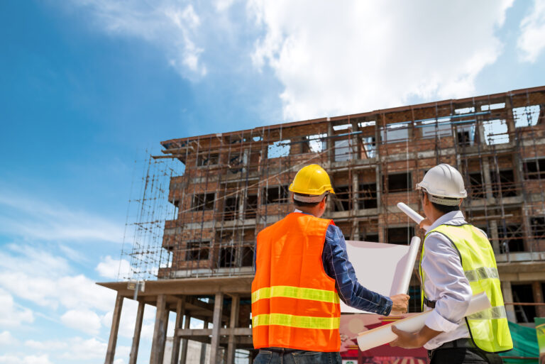 Two site engineers in safety vests and hard hats analyzing a digital-ready blueprint against a multi-story building frame, illustrating smart construction trends 2026 in project management.