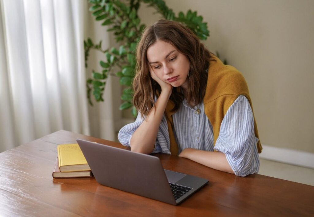 A young woman sitting at a desk at home looking frustrated while reviewing exam results on her laptop
