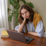 A young woman sitting at a desk at home looking frustrated while reviewing exam results on her laptop
