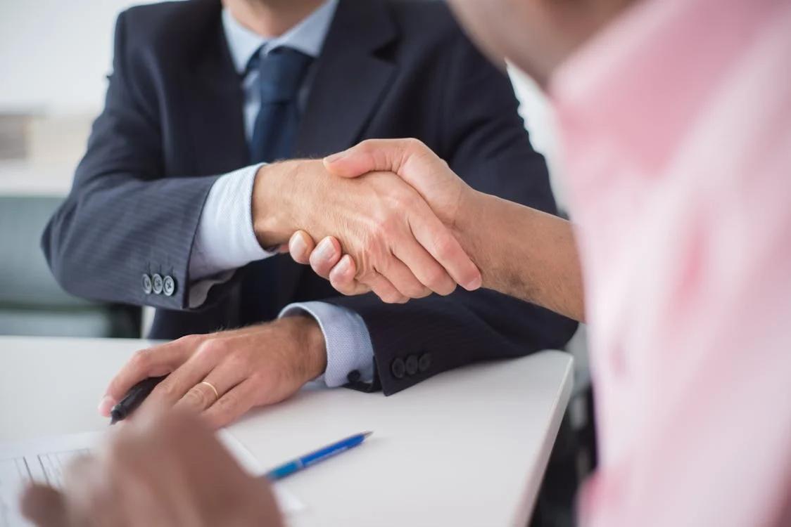 two people sitting together by a white table, shaking hands after signing a form