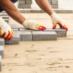 A close-up view of a professional's gloved hands meticulously laying grey concrete interlocking pavers onto a prepared sand base, illustrating the precision of professional landscaping and hardscape installation.