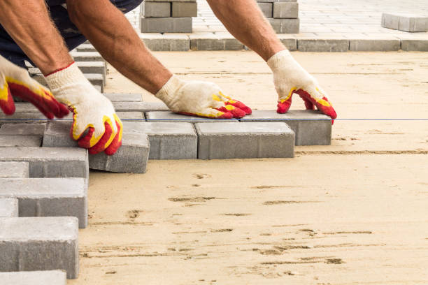 A close-up view of a professional's gloved hands meticulously laying grey concrete interlocking pavers onto a prepared sand base, illustrating the precision of professional landscaping and hardscape installation.