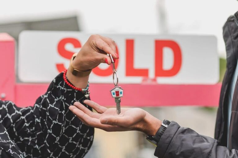 a person handing over the keys to a house to another person in front of the “sold” sign