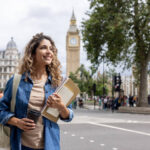 A smiling young female student carrying notebooks and a coffee cup walking in central London with Big Ben in the background, representing the experience of study in UK for international students.