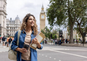 A smiling young female student carrying notebooks and a coffee cup walking in central London with Big Ben in the background, representing the experience of study in UK for international students.