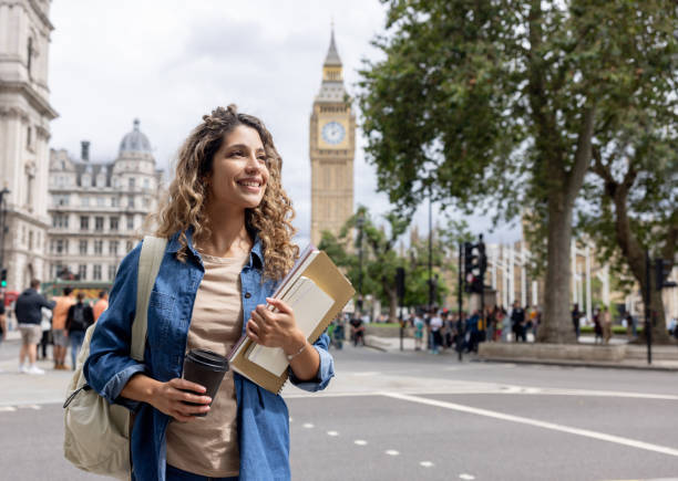 A smiling young female student carrying notebooks and a coffee cup walking in central London with Big Ben in the background, representing the experience of study in UK for international students.