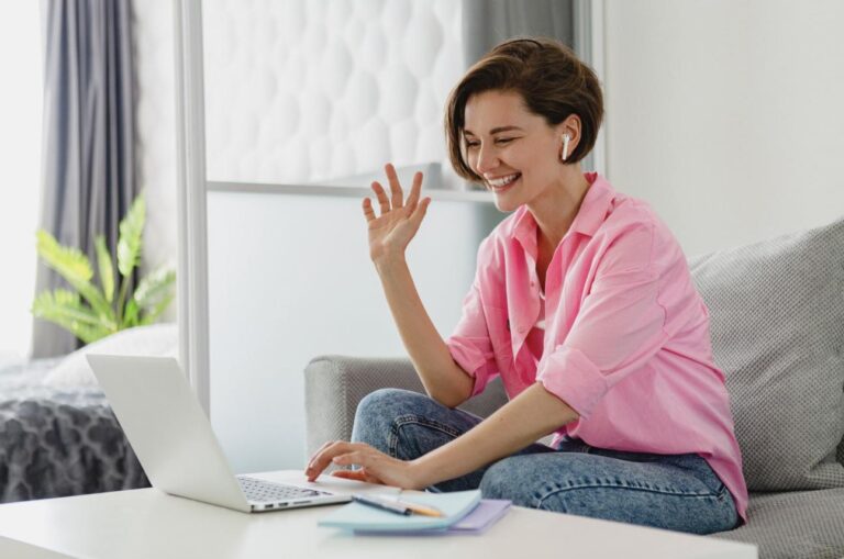 A woman sitting on her sofa at home, smiling and waving during a private online therapy session