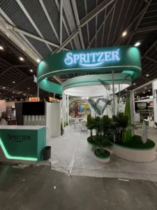 Spritzer exhibition booth with green curved signage, illuminated logo, plants, and seating area inside a convention hall.