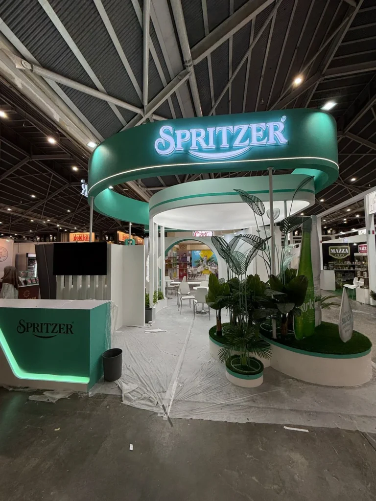 Spritzer exhibition booth with green curved signage, illuminated logo, plants, and seating area inside a convention hall.