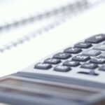 Close-up image of a person pressing buttons on a calculator beside a notebook and stacked coins on a desk.