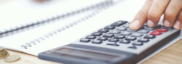 Close-up image of a person pressing buttons on a calculator beside a notebook and stacked coins on a desk.