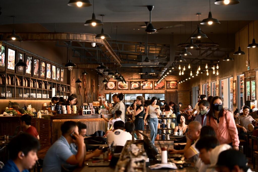 A crowded restaurant interior with diners seated at wooden tables under pendant lights, staff moving through the floor, and illuminated menu boards visible along the walls.