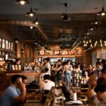 A crowded restaurant interior with diners seated at wooden tables under pendant lights, staff moving through the floor, and illuminated menu boards visible along the walls.