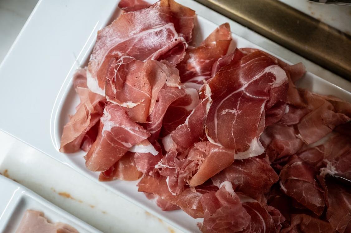 A close-up of thinly sliced cured deli meat arranged on a white serving tray with tongs visible in the background.