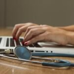 An image of a person typing on a laptop with a stethoscope next to them