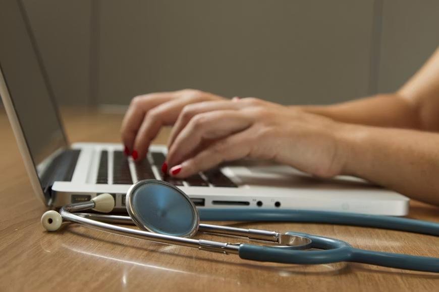 An image of a person typing on a laptop with a stethoscope next to them
