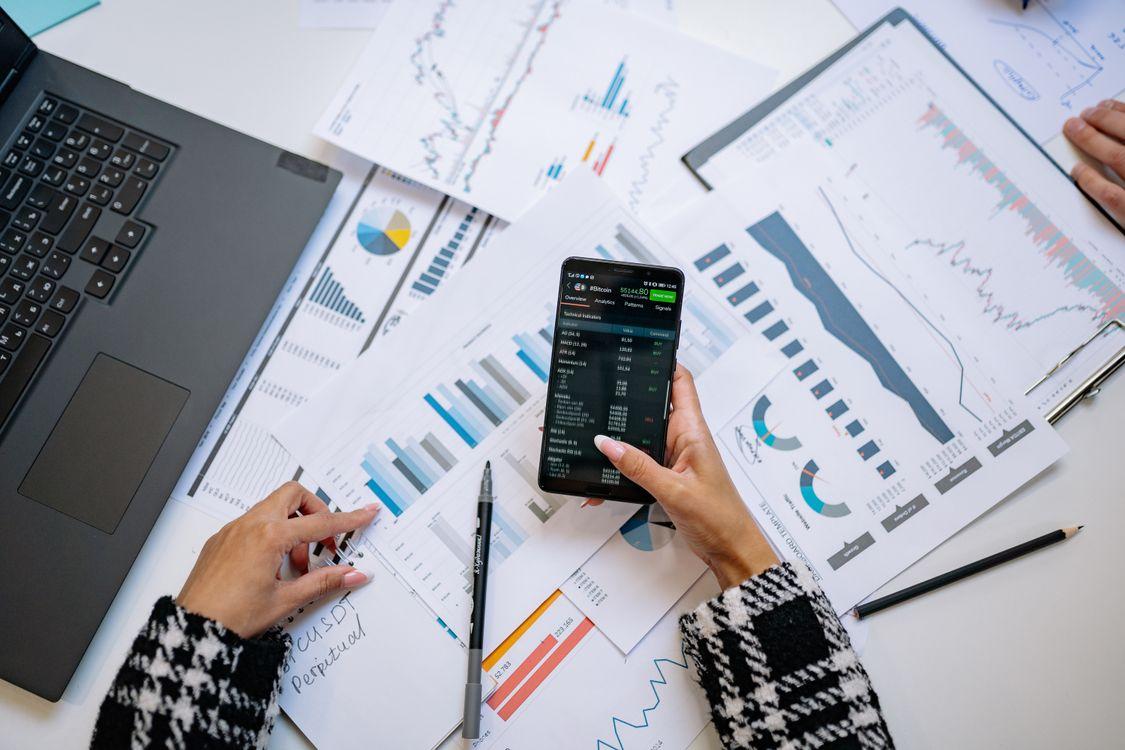 Person holding smartphone with laptop and financial documents on table