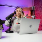 Female content creator wearing headphones recording audio with a microphone and laptop in a modern studio setup