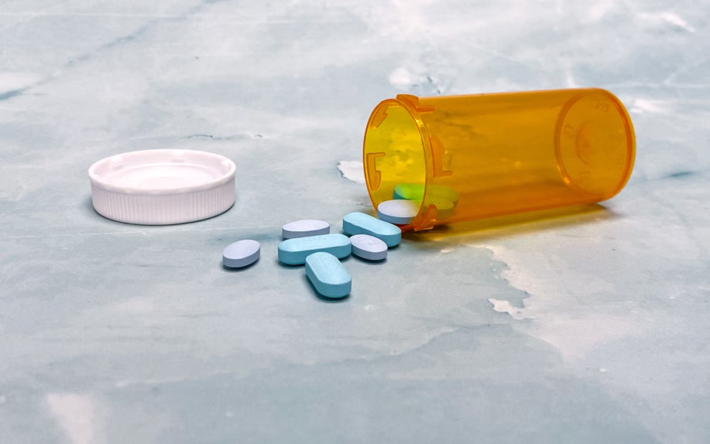 Open orange prescription bottle with blue and white tablets spilled onto a gray surface beside its cap.