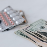 Blister packs of pills placed beside a stack of U.S. dollar bills on a white surface.