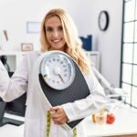 beautiful-blonde-nutritionist-woman-holding-weight-machine-balance-weight-loss-pointing-thumb-up-side-smiling-happy-with-open-mouth-2-1024x683
