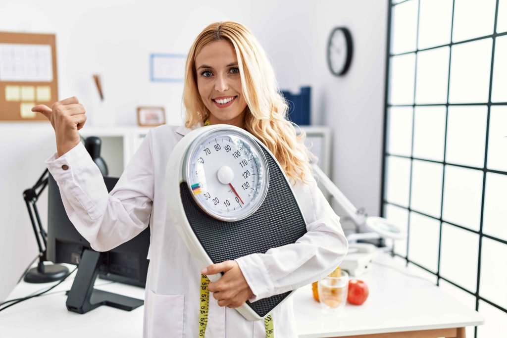 beautiful-blonde-nutritionist-woman-holding-weight-machine-balance-weight-loss-pointing-thumb-up-side-smiling-happy-with-open-mouth-2-1024x683