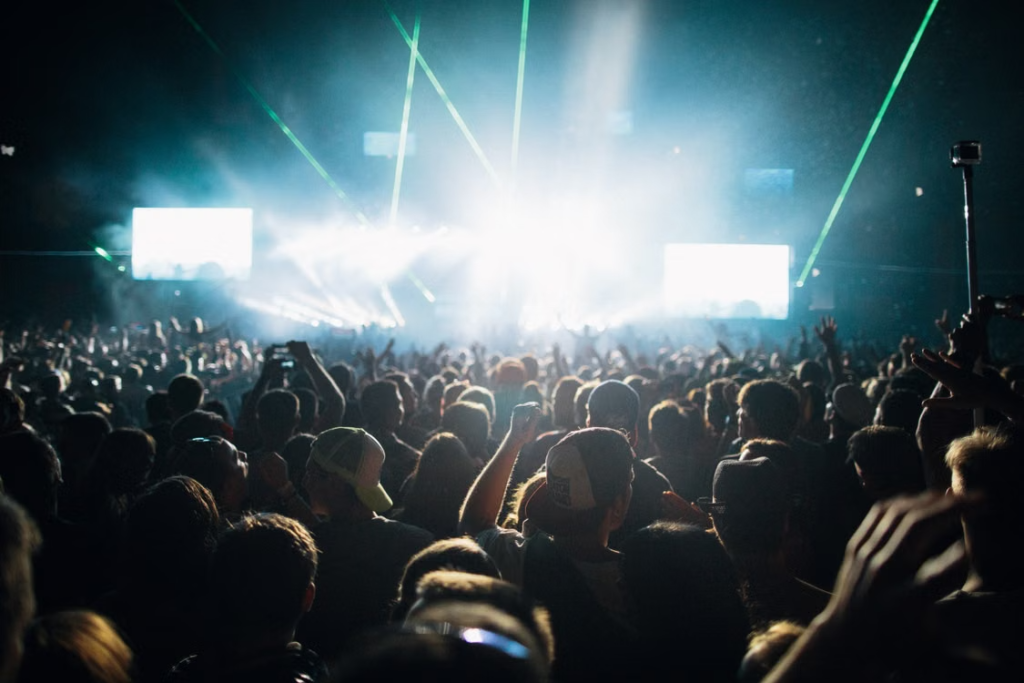 A crowd with raised hands facing a brightly lit stage with green laser beams and large screens.